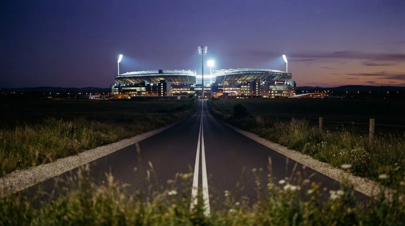 Ein Flutlichtstadion erhebt sich bei Dämmerung in der Ferne hinter einer leeren Landstraße, die Lichter sind eingeschaltet.