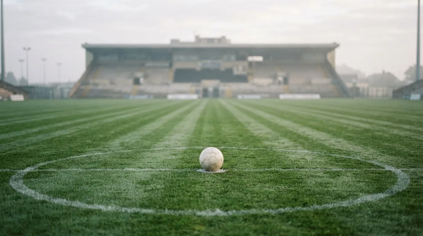Ein Fußball liegt auf dem Mittelkreis eines leeren Rasenplatzes am frühen Morgen, im Hintergrund eine ferne Tribüne im Dunst.