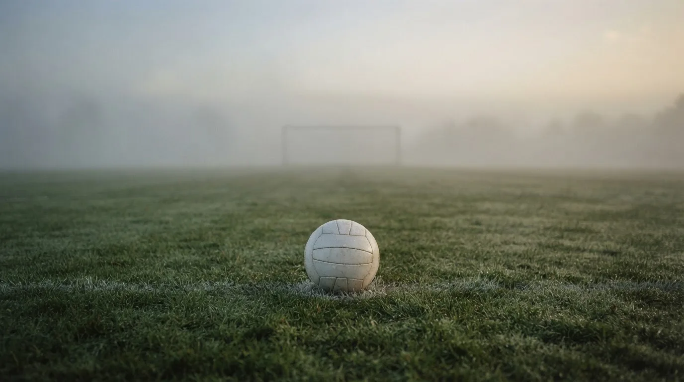 Ein Fußball liegt auf dem Anstoßpunkt eines leeren Rasenplatzes im Morgennebel.