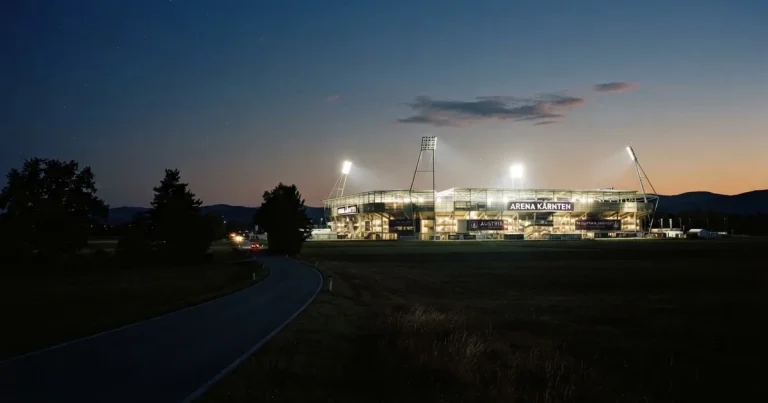 Ein Stadion unter Flutlicht aus großer Entfernung gesehen, davor eine dunkle Landstraße und einzelne Bäume in der Abenddämmerung.